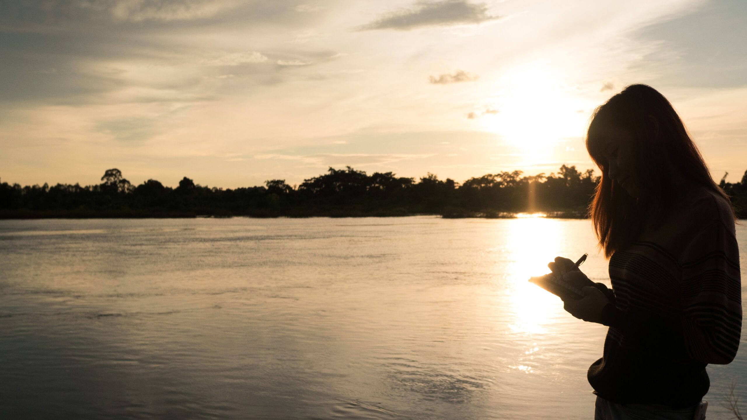 Young woman with pen writing on notebook at riverside in the evening.